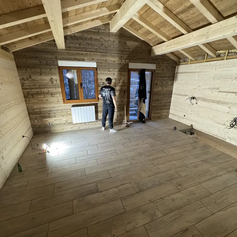 Grande pièce sous charpente apparente, murs en lambris, sol en carrelage imitation bois, un compagnon de dos près de la fenêtre