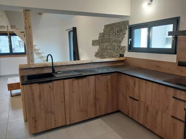 Cuisine en angle aux façades bois et plan de travail noir, pan de mur en pierre de parement, escalier au fond