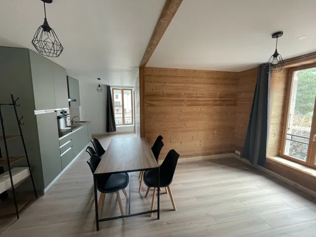 Cuisine aux façades vertes ouverte sur un séjour, mur en lambris, table et chaises noires