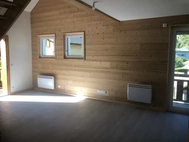 Pièce sous rampant avec un mur entier en lambris clair, deux petites fenêtres, radiateurs et prises posés